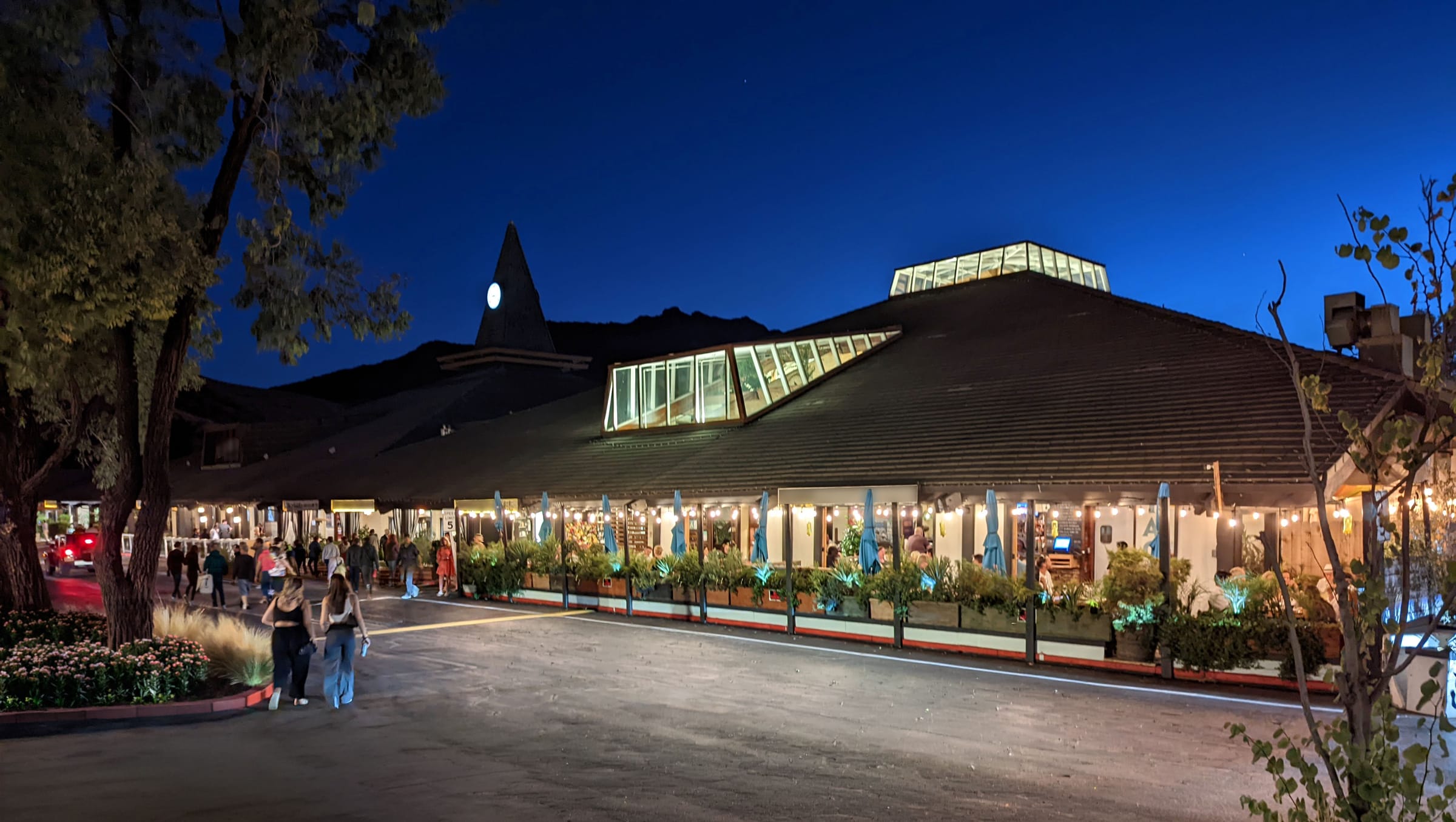 A commercial market square at dusk — string lights on, shops open, people walking the promenade